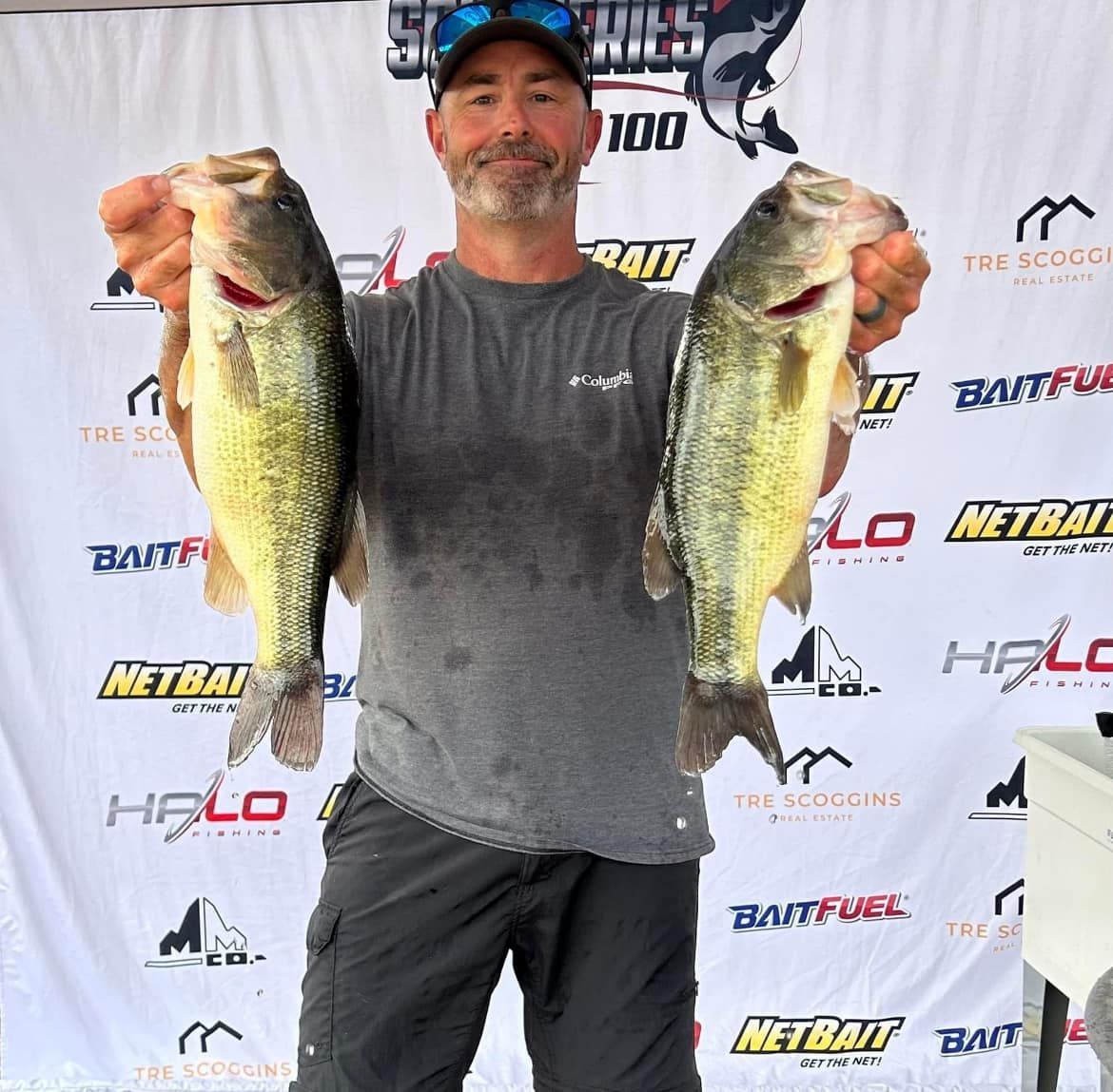 Angler holding two large bass in front of a sponsor backdrop after a fishing event