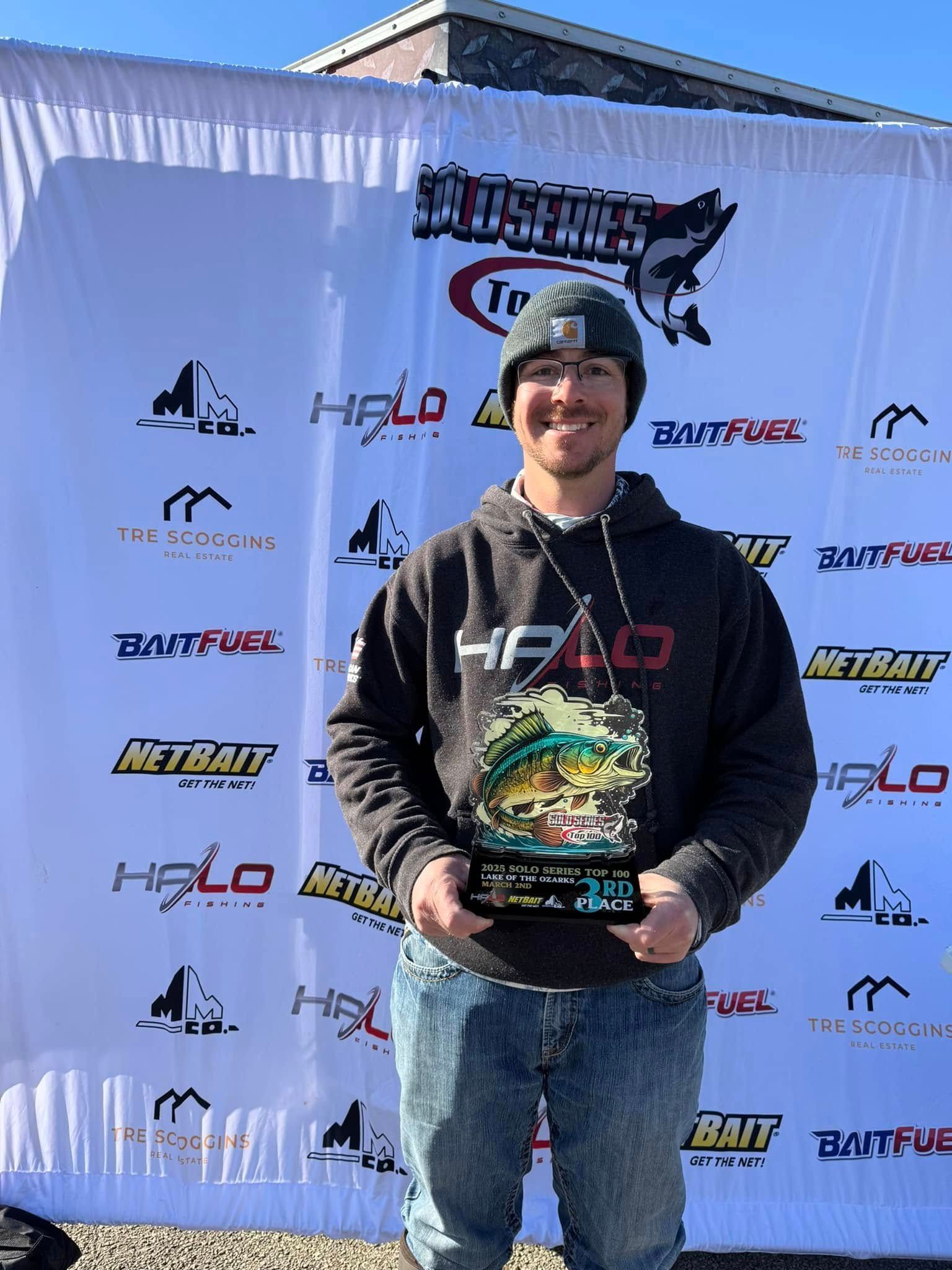 Person holding a trophy in front of a sponsor backdrop, wearing a black hoodie and beanie, smiling outdoors.