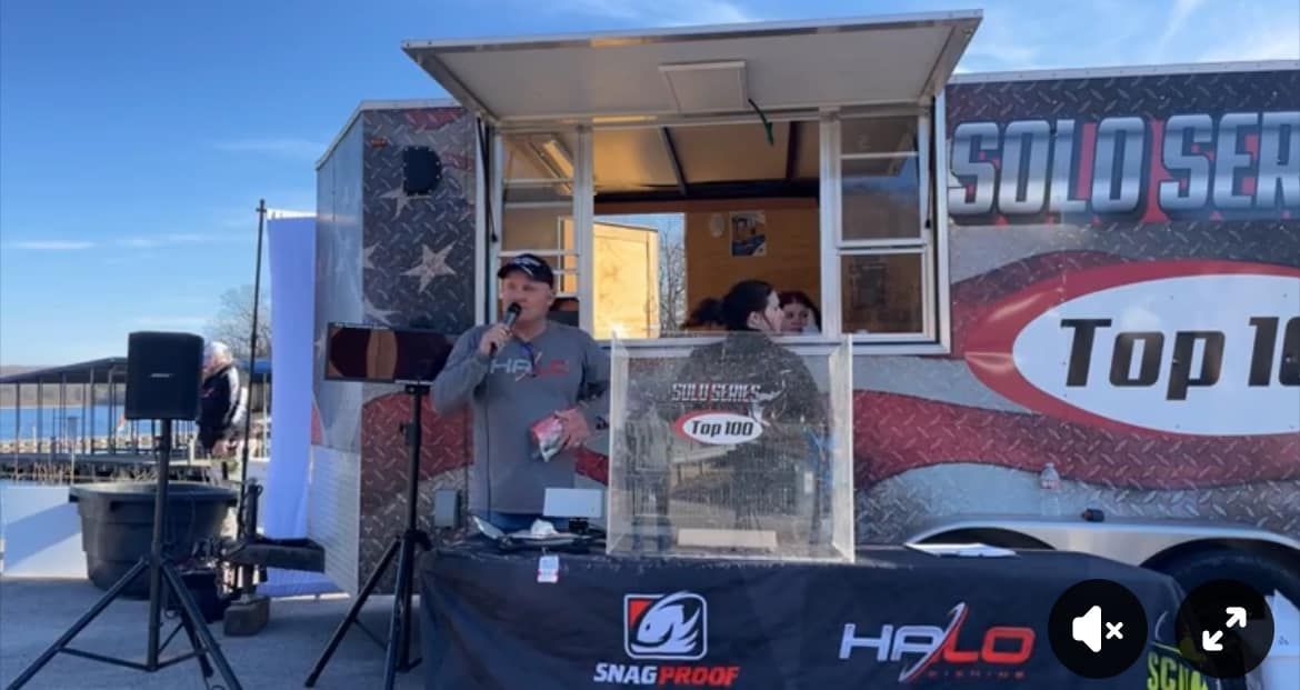 Outdoor event booth with a speaker at a microphone beside a “Top Fuel” trailer display.