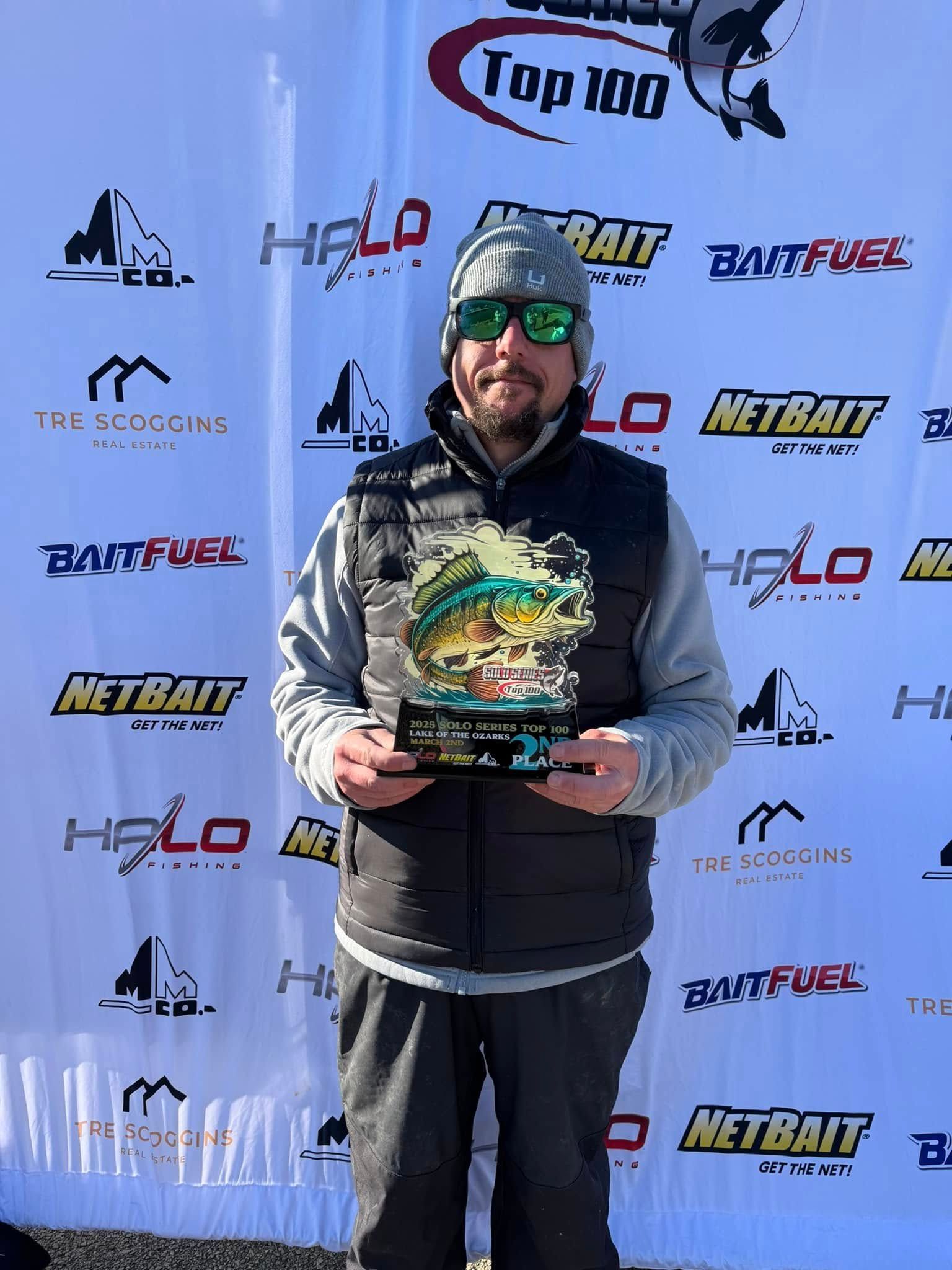 Man in sunglasses holding a trophy in front of sponsor banners at an outdoor event