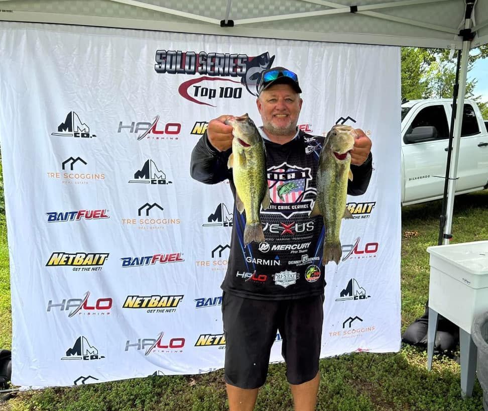 Man holding two fish at an outdoor fishing tournament awards backdrop, smiling in a black jersey and cap