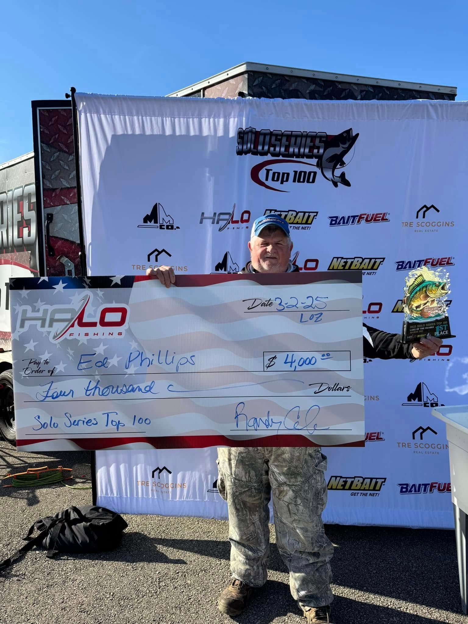 Person in camouflage holding a large check in front of a sponsor backdrop outdoors