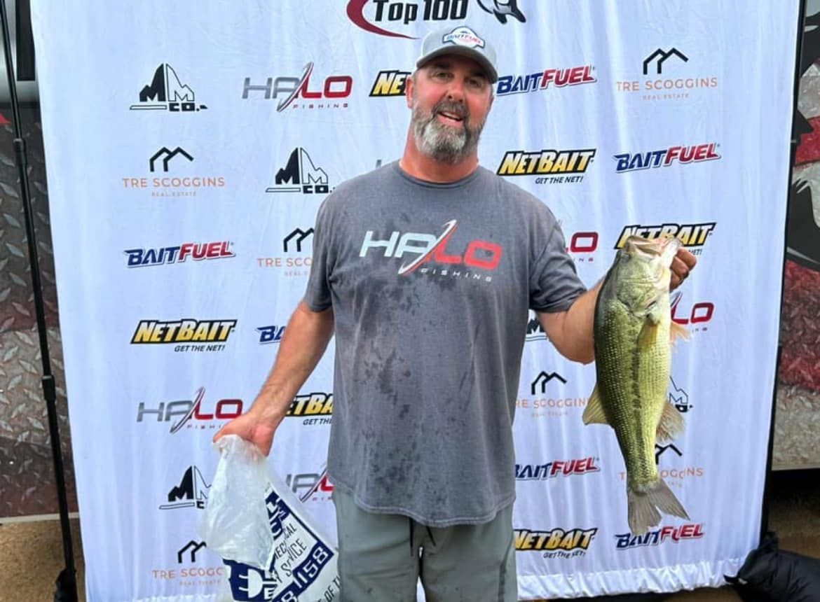 Angler smiling and holding a large bass at a fishing tournament backdrop.