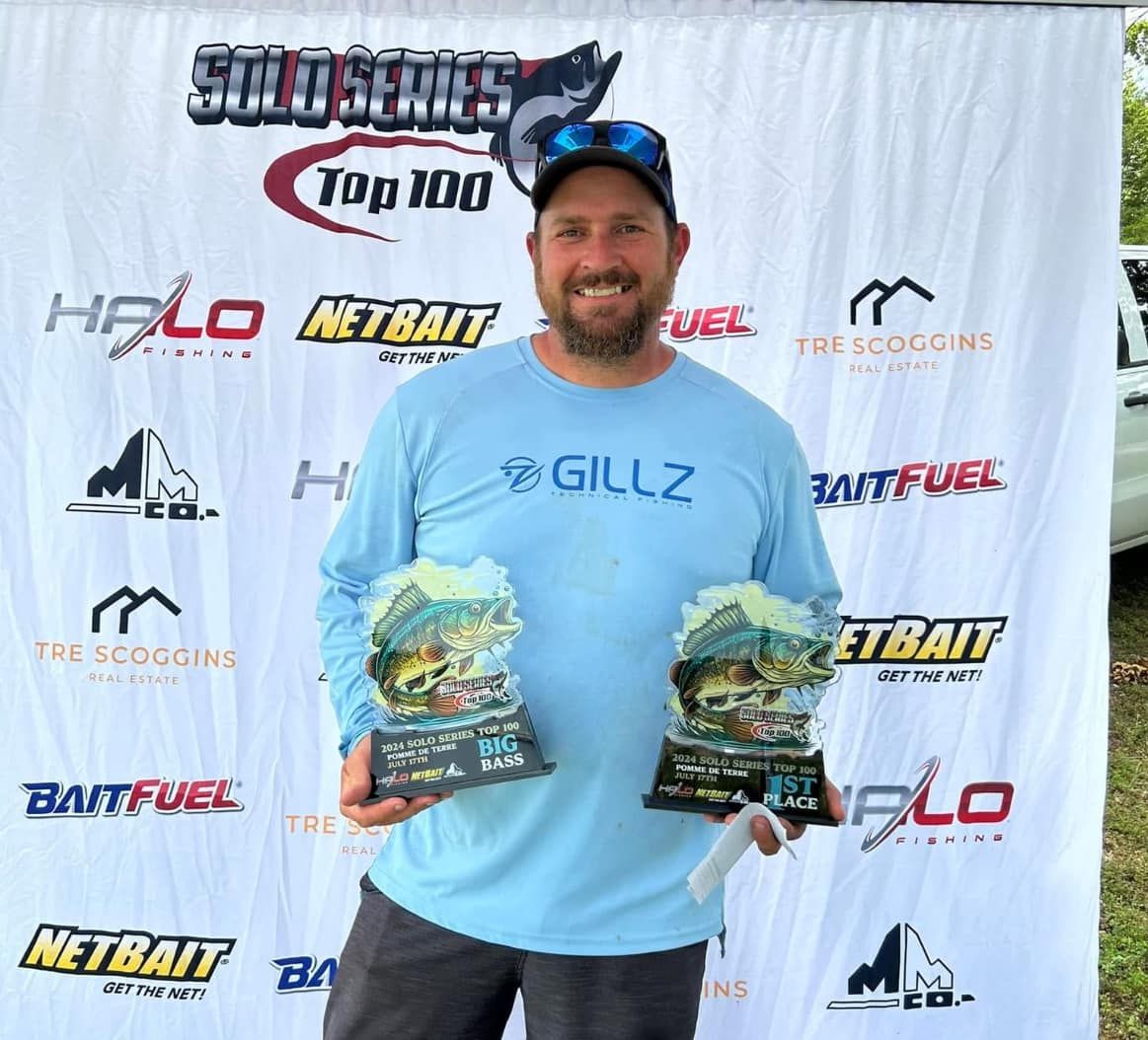 Man in blue shirt holding two fish trophies in front of sponsor backdrop