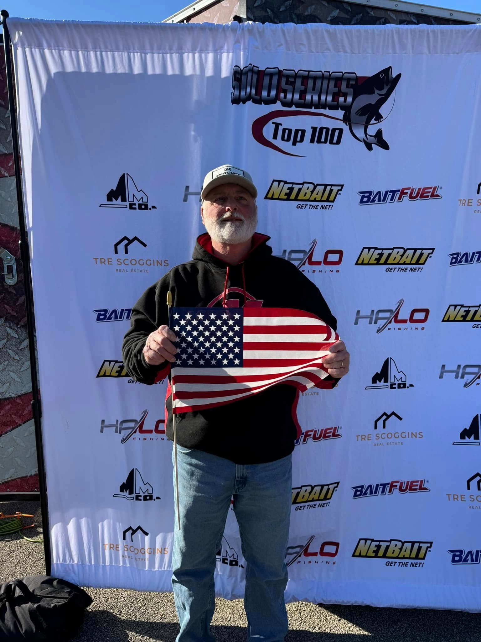 Man holding a U.S. flag plaque in front of a sponsor banner outdoors