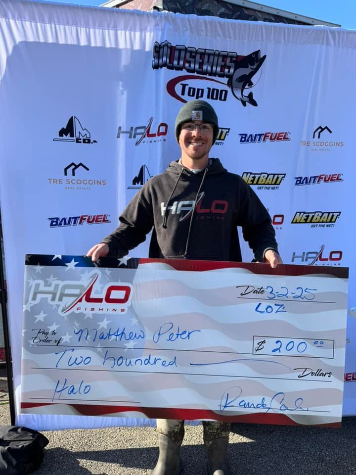 Person holding a large check at a fishing event, standing in front of sponsor banners.