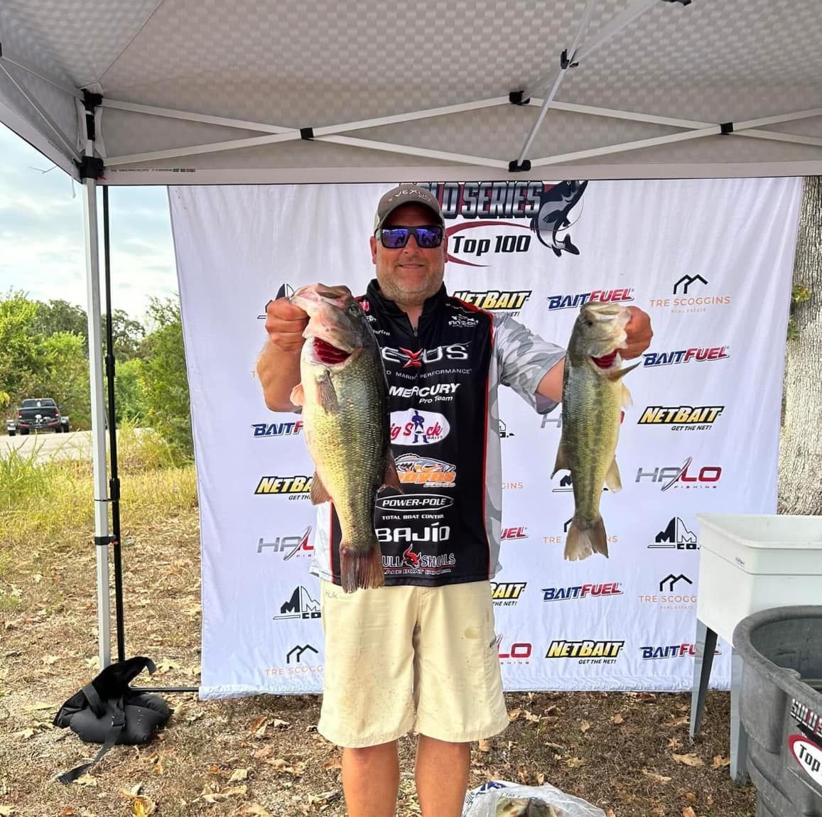 Man under a canopy holding two large bass fish at a fishing tournament backdrop