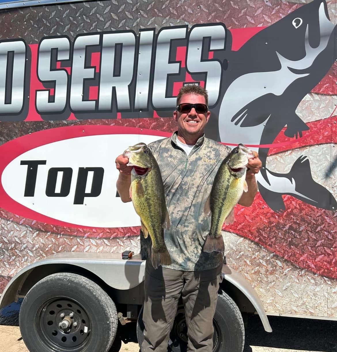 Man holding two fish in front of a fishing series trailer mural, wearing sunglasses and camo shirt.