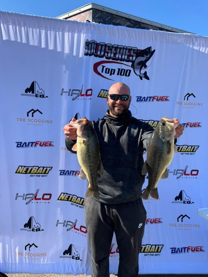 Man holding two large fish in front of a sponsor backdrop at an outdoor fishing event