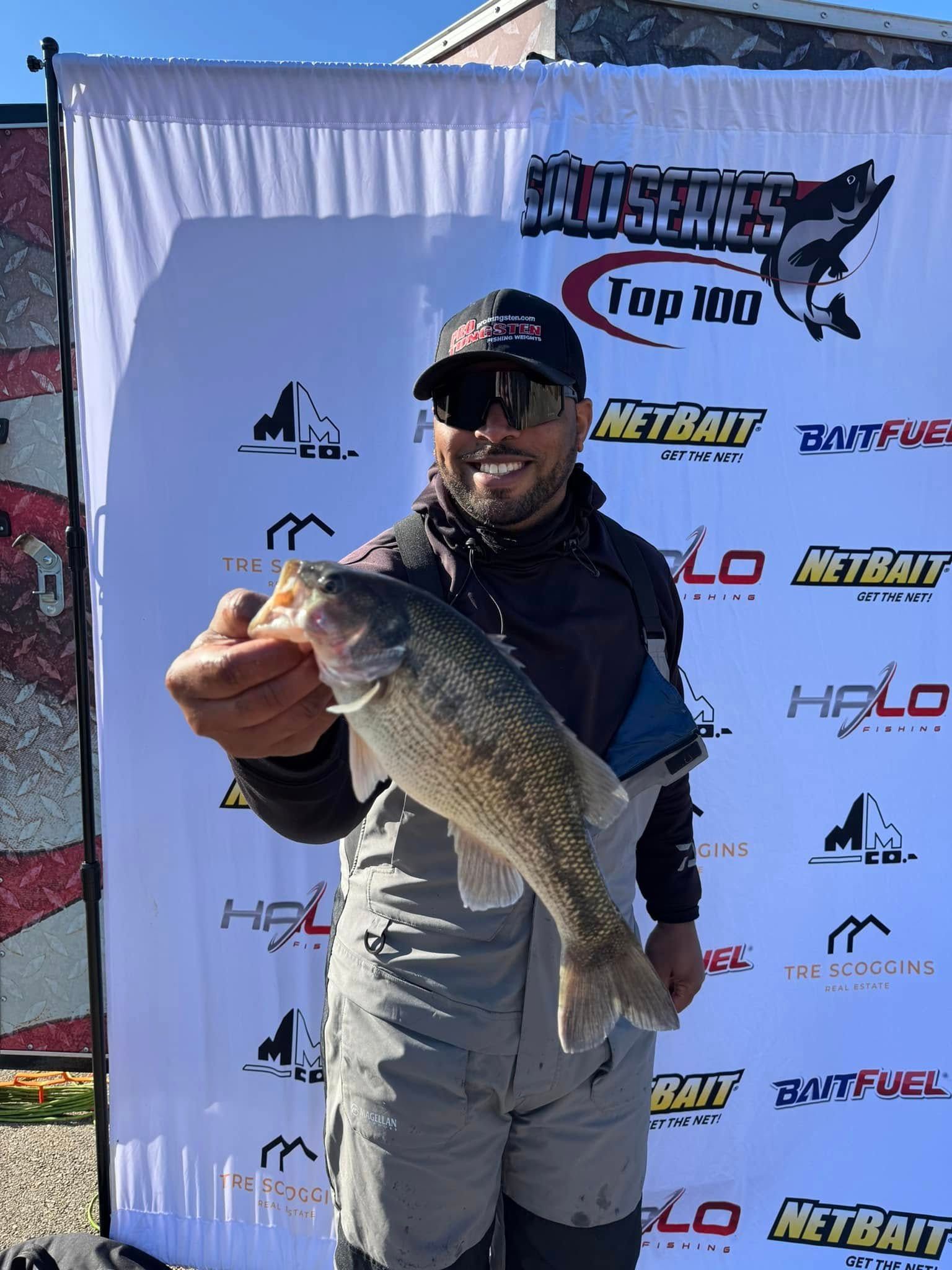 Angler posing with a large fish in front of a sponsor banner at a fishing event