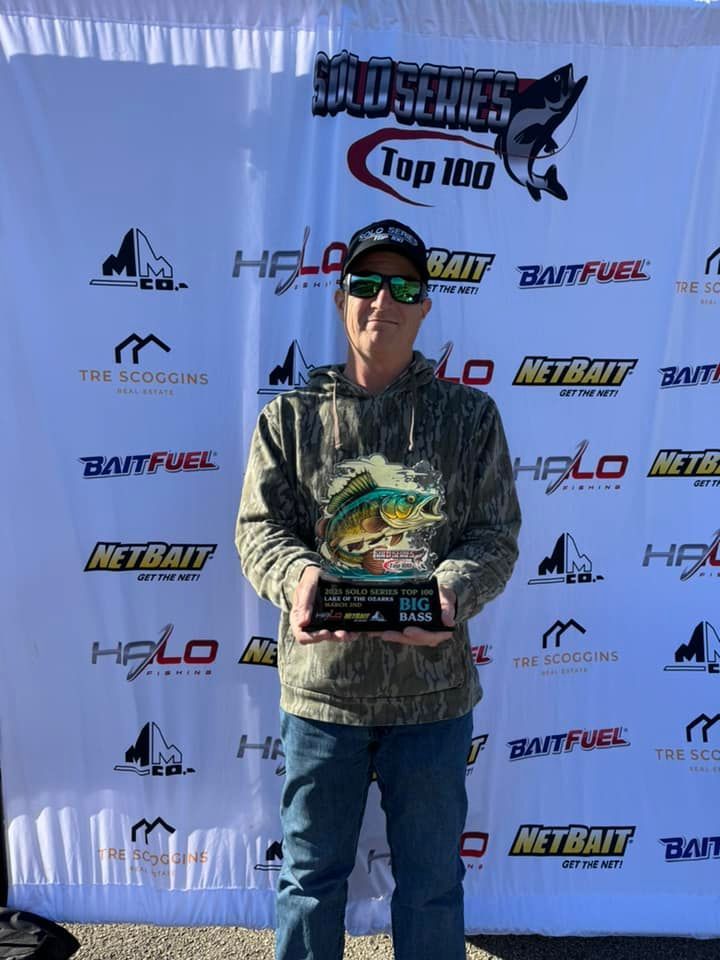 Person holding a trophy in front of a sponsor banner at an outdoor fishing event