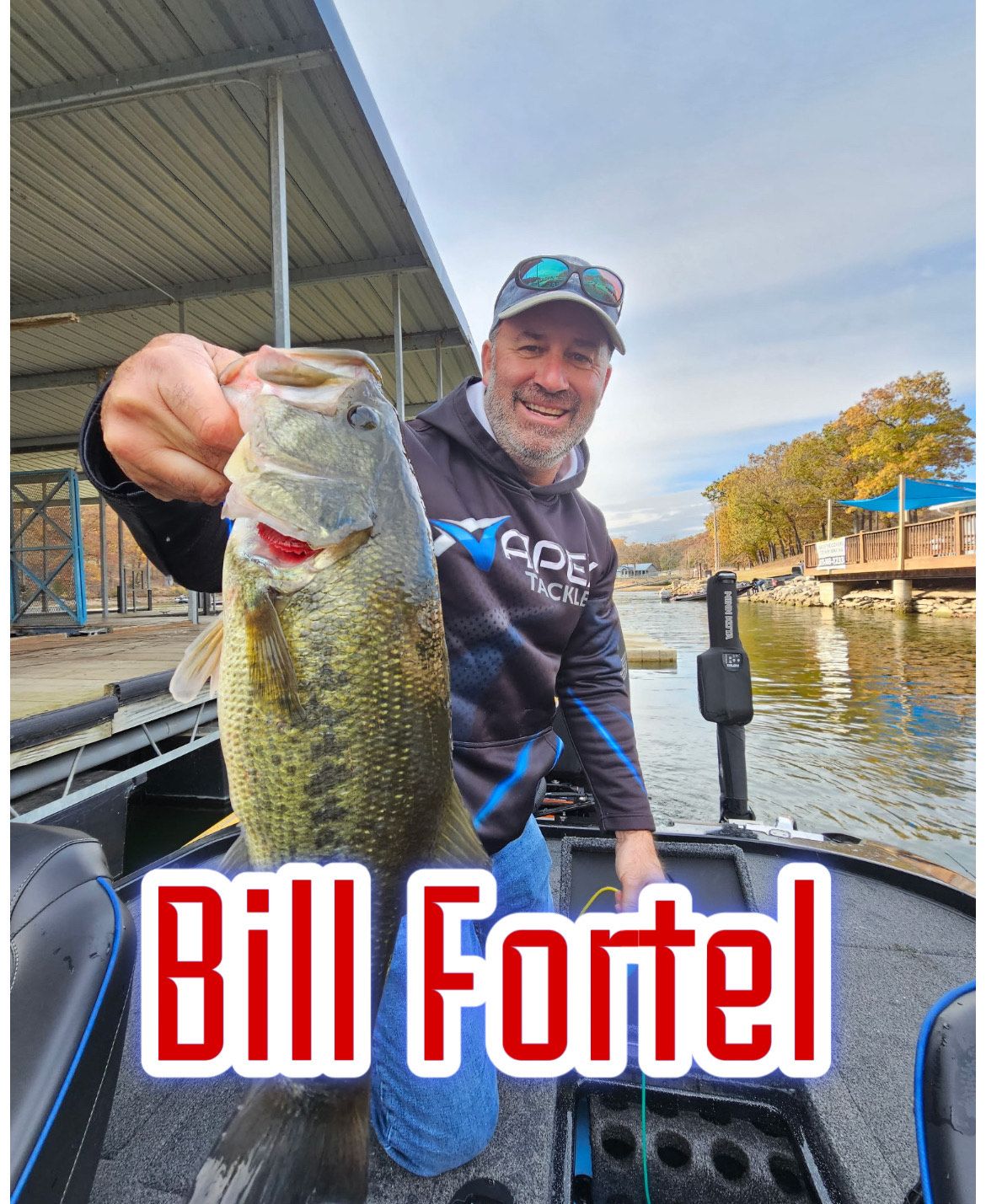 Man holding a largemouth bass on a boat at a lakeside dock, with “Bill Fortel” text overlay.