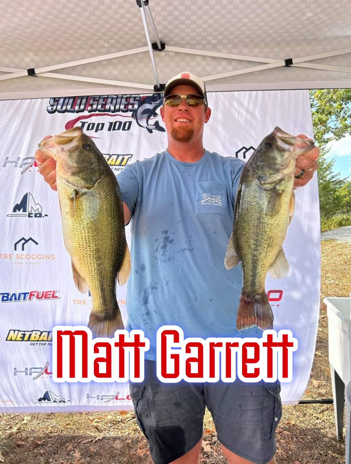 Angler holding two large bass under a canopy, with a tournament banner behind him.