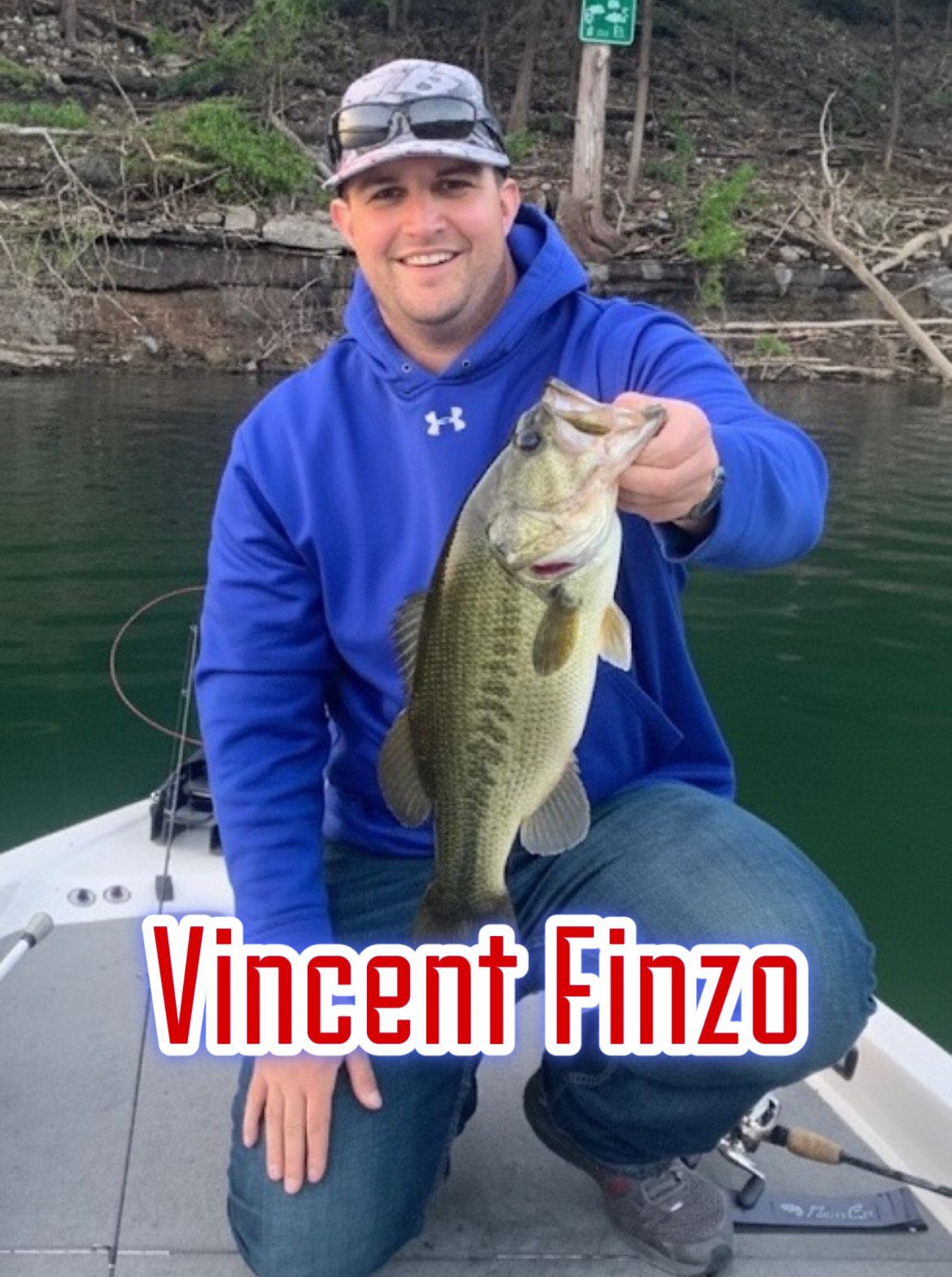 Man in blue hoodie kneeling on a boat, holding a largemouth bass on a lake, with “Vincent Finzo” text.
