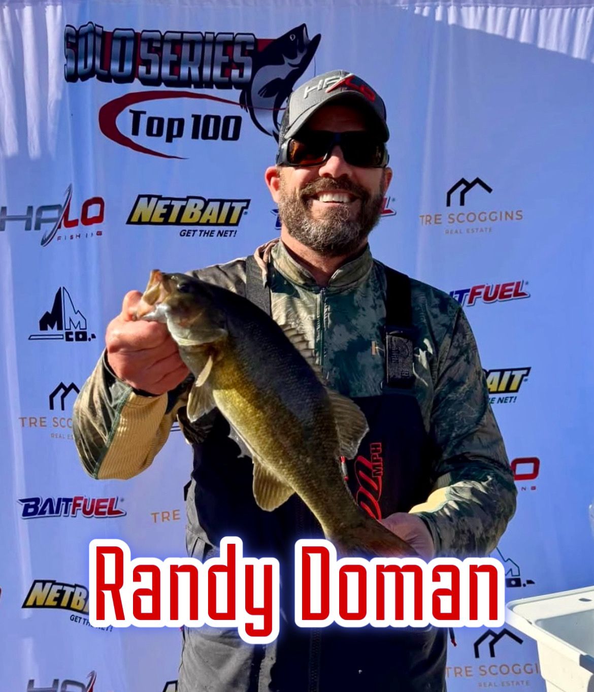 Angler holding a largemouth bass at a tournament, with sponsor logos and name Randy Doman in the background.