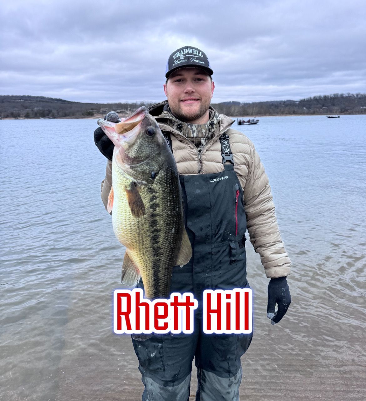 Angler holding a large bass in shallow water with cloudy sky and the name “Rhett Hill” overlaid.