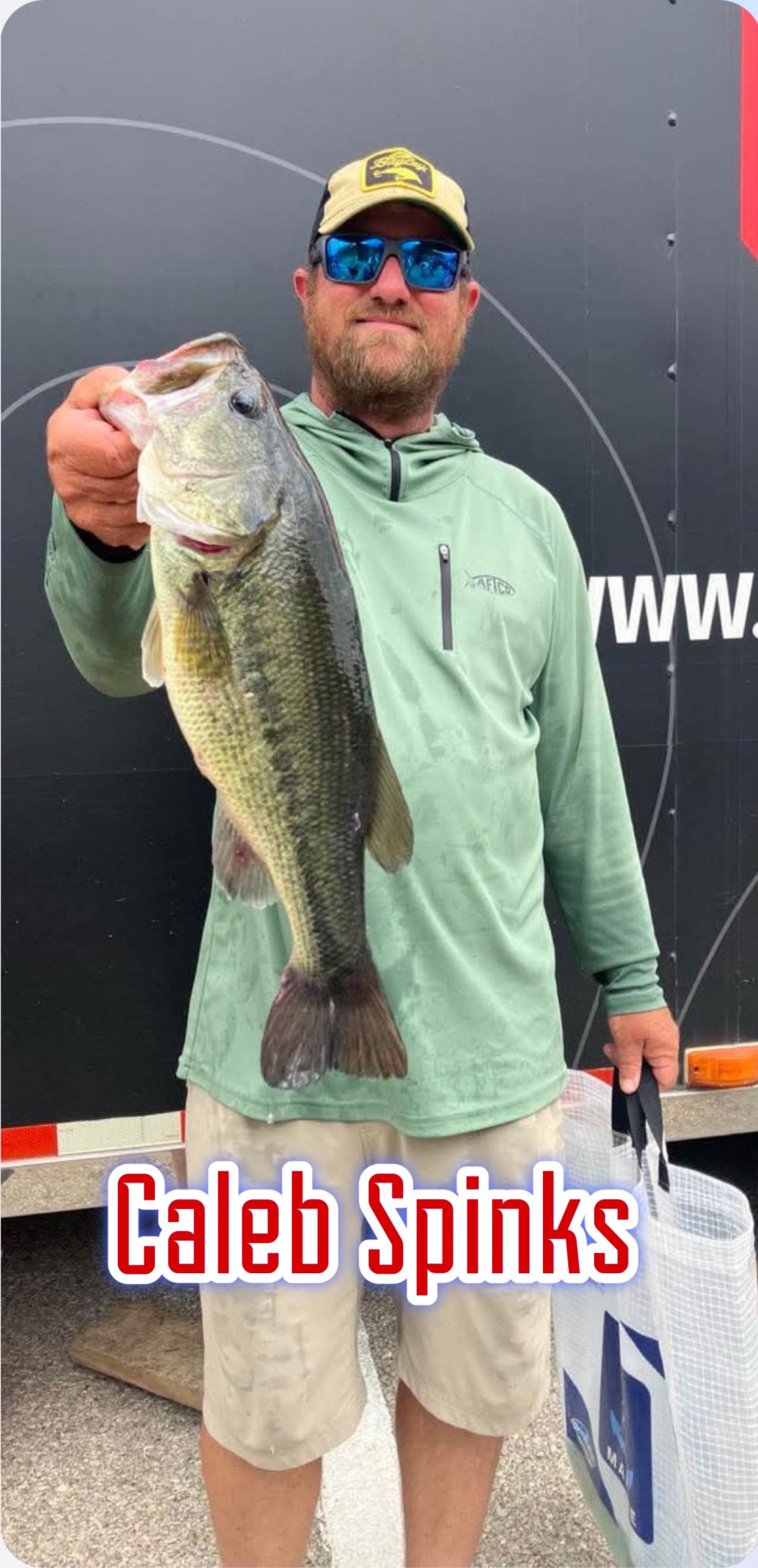 Man holding a large fish on a stage, wearing a cap, sunglasses, and green shirt, with “Caleb Spinks” text below