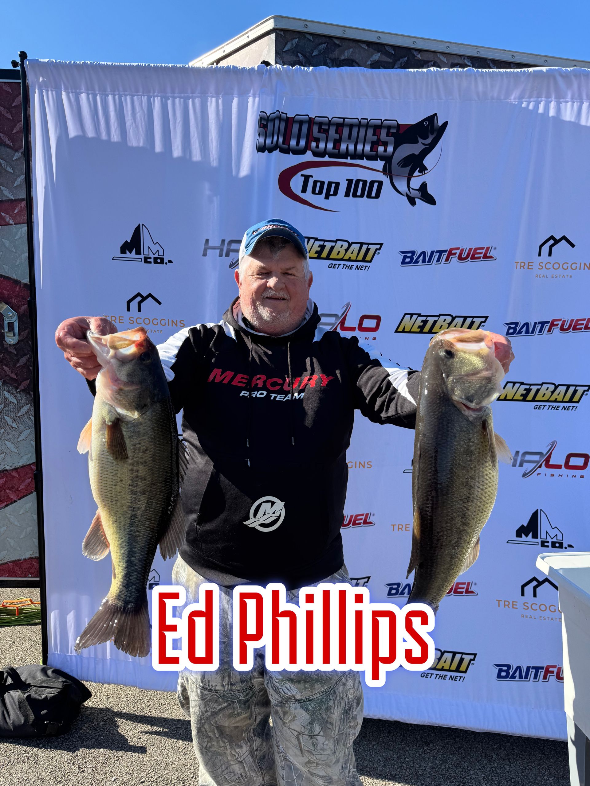Man holding two large fish in front of a sponsor backdrop, with “Ed Phillips” text below