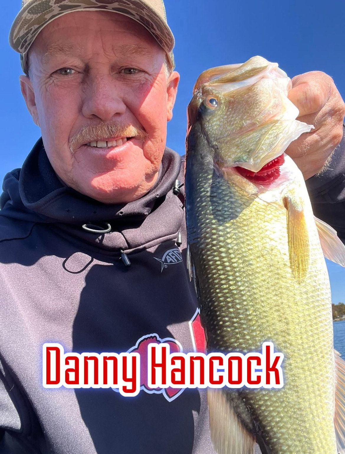 Man holding a largemouth bass against a blue sky, with “Danny Hancock” text at the bottom