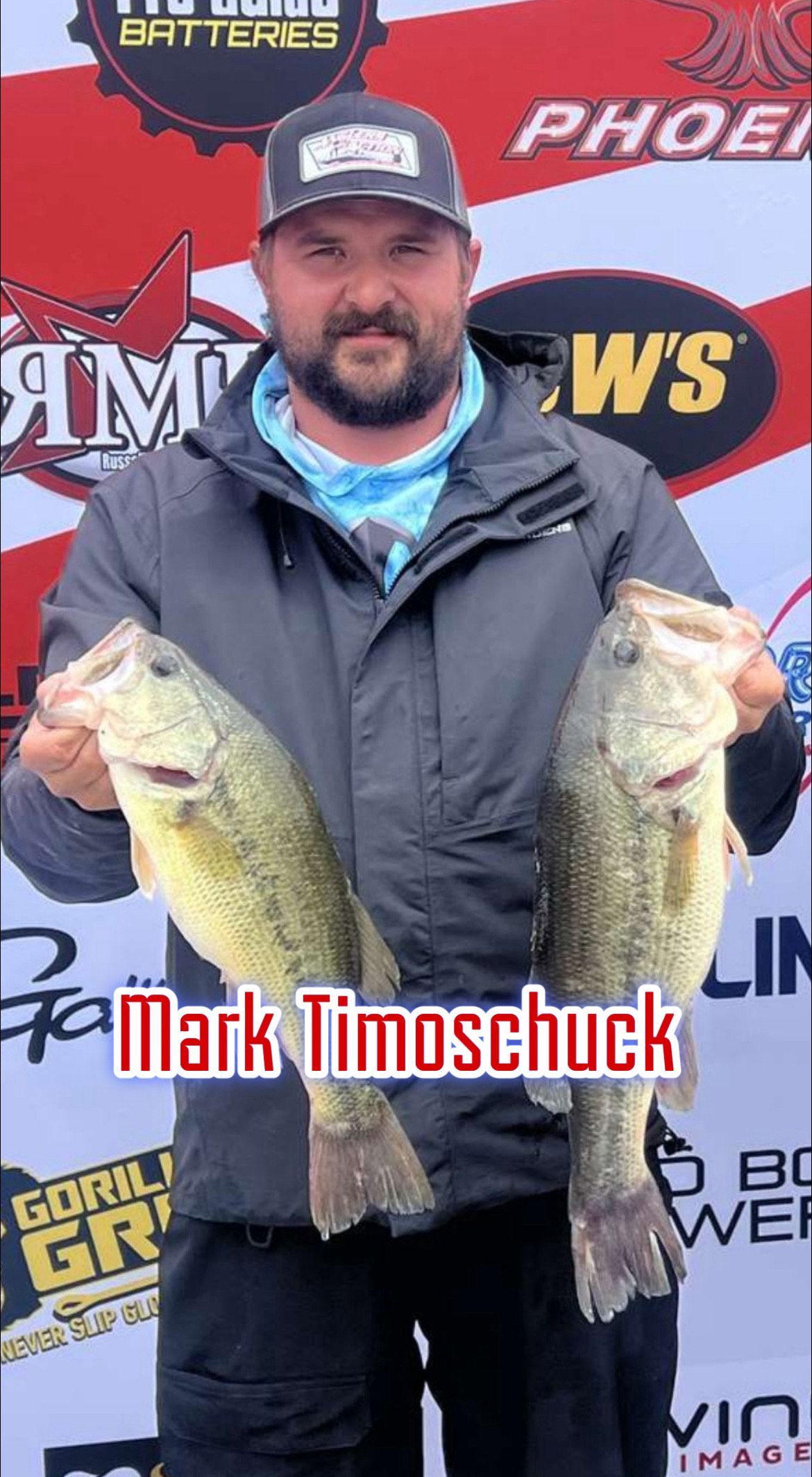 Man holding two large fish at a fishing event, standing in front of sponsor banners.