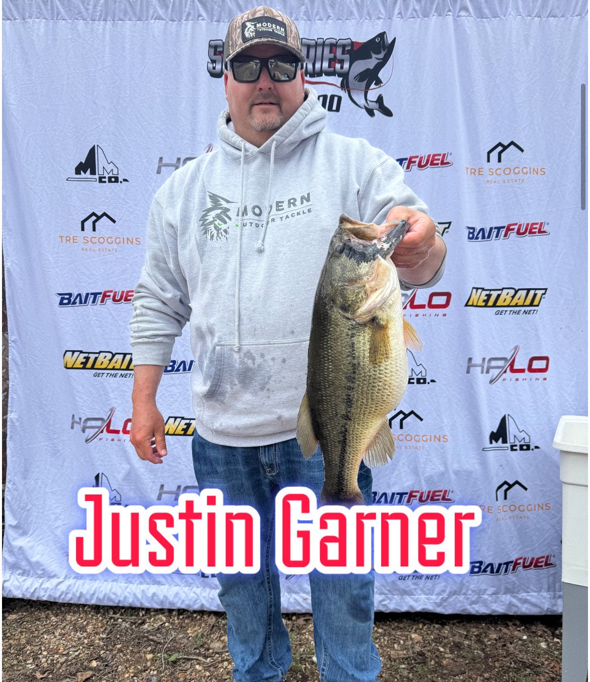 Man posing with a large fish at a fishing event backdrop, with “Justin Garner” text at bottom.