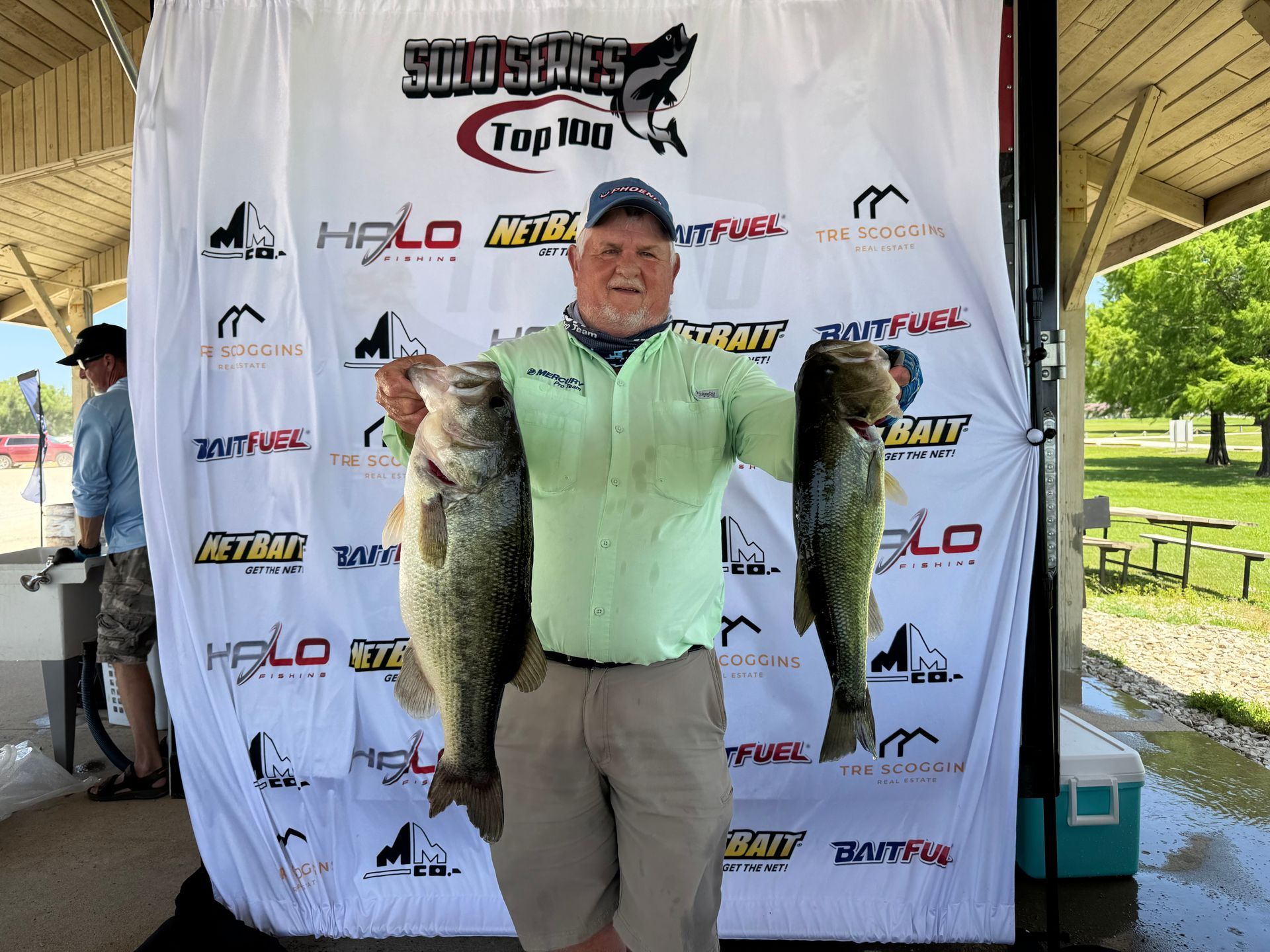 Man holding two large fish at a fishing tournament podium with sponsor backdrop