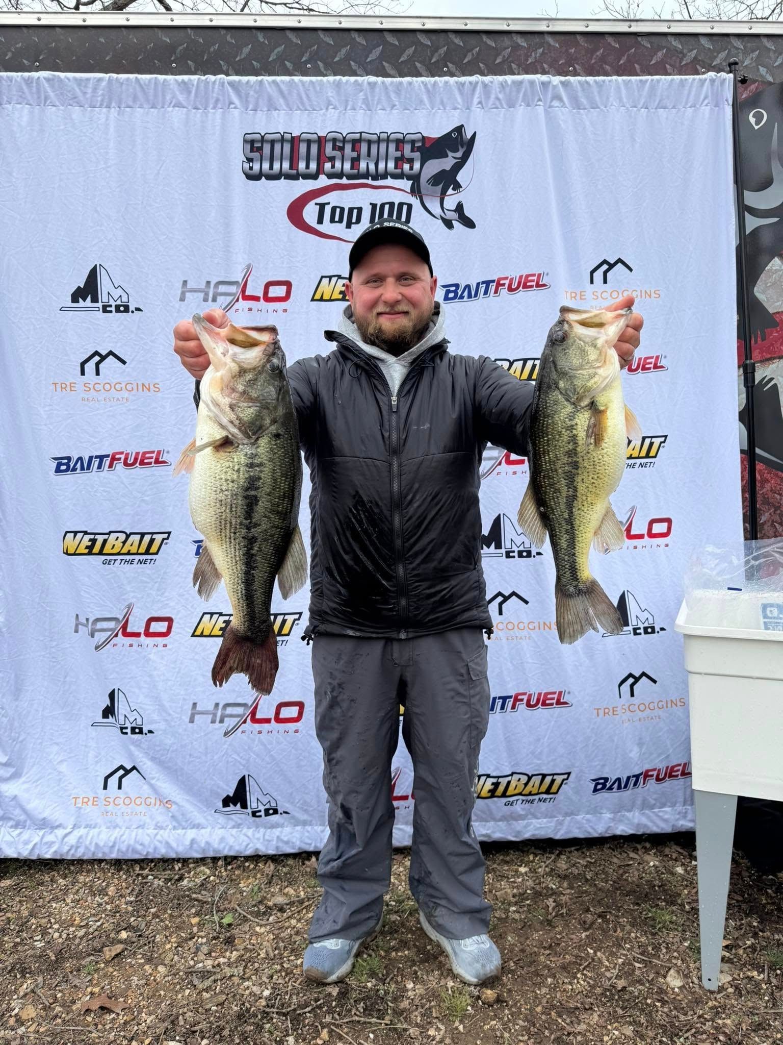 Man holding two large fish at an outdoor fishing event backdrop