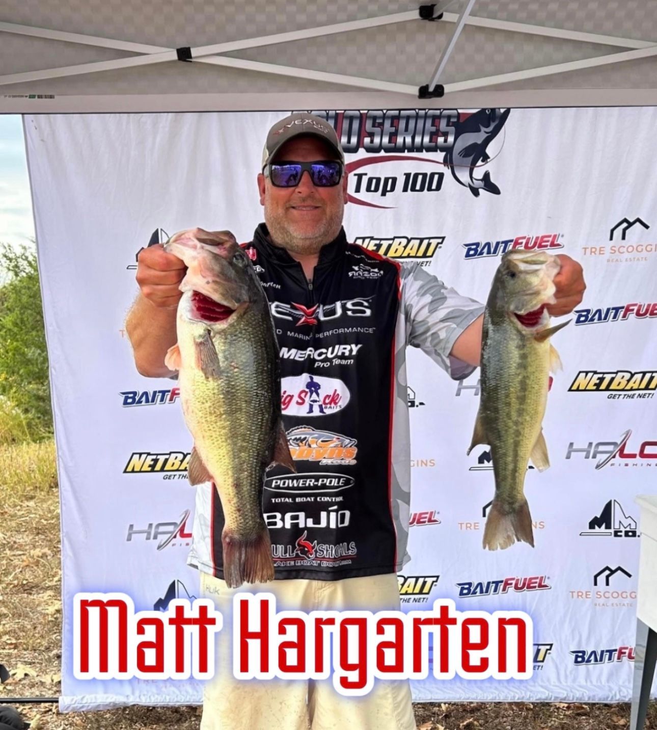 Angler holding two largemouth bass at a fishing tournament, with sponsor banner behind him.