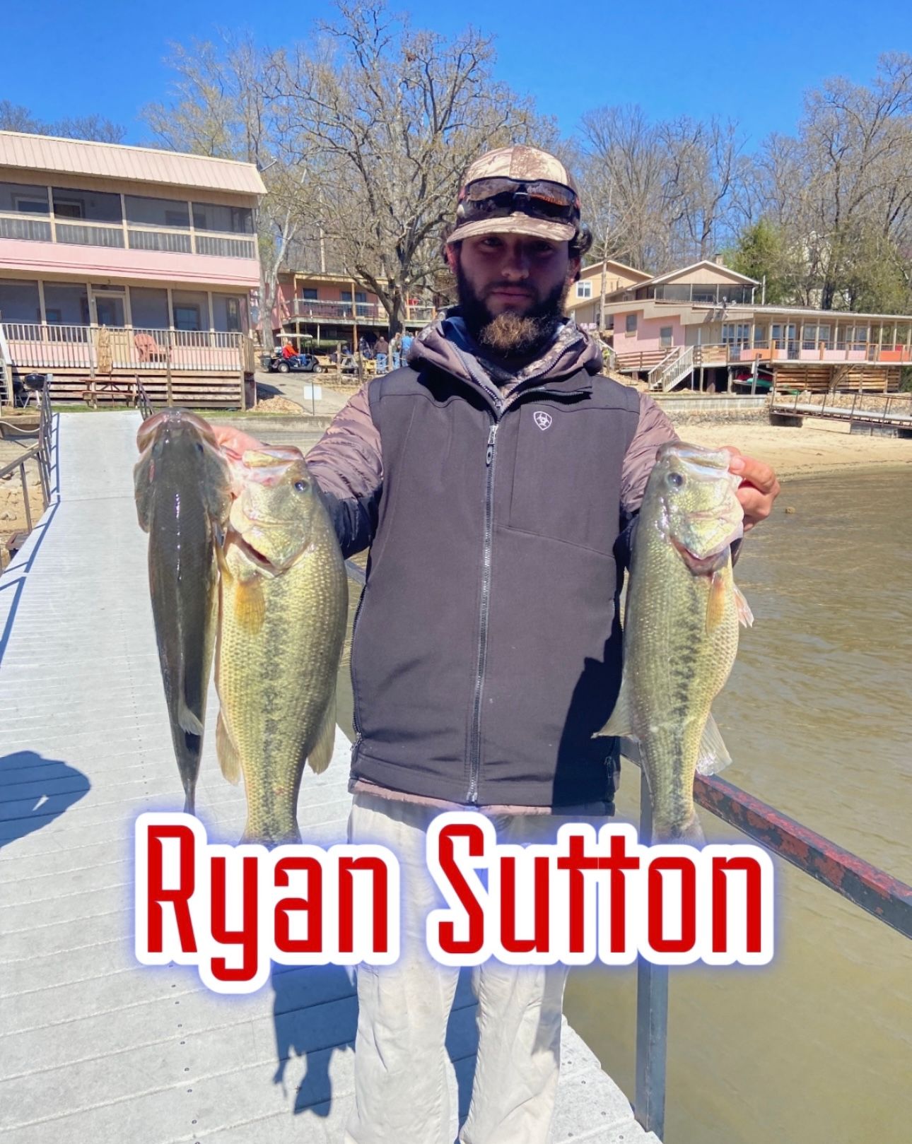 Man holding two large fish on a dock with boats and cabins in the background; text reads “Ryan Sutton”