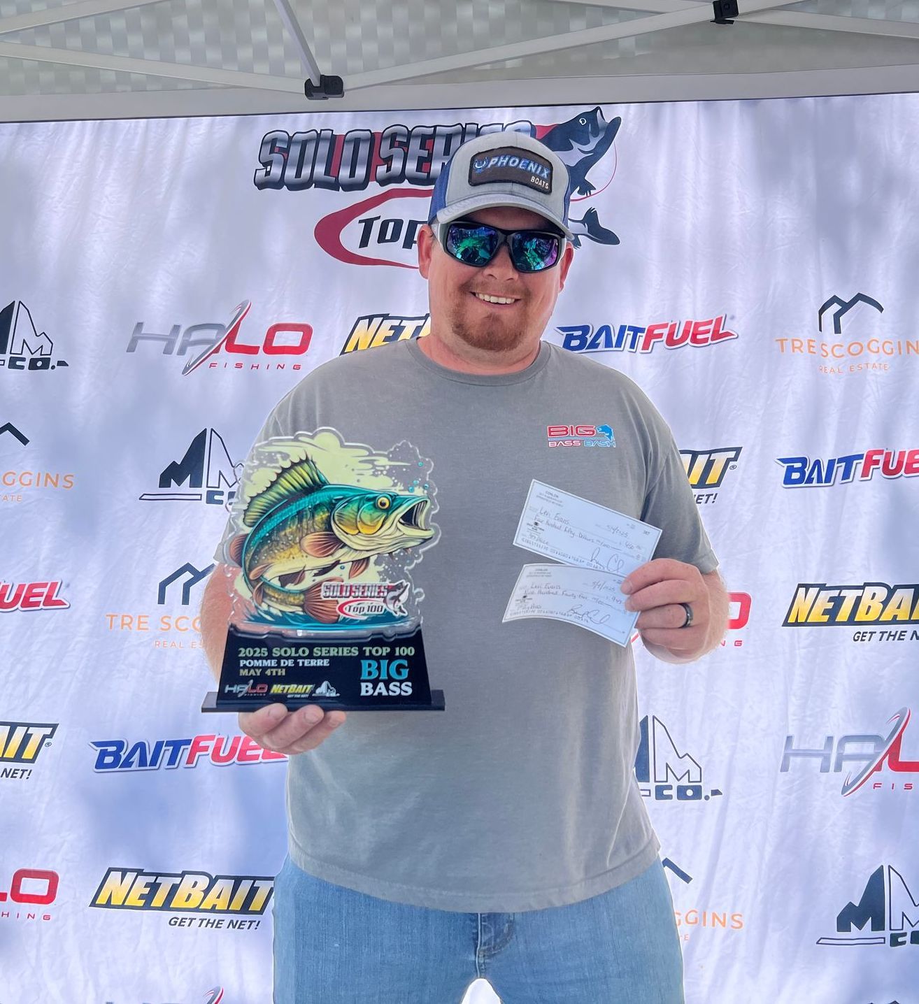 Man holding a trophy and checks at a bass fishing event podium, wearing sunglasses and a cap.