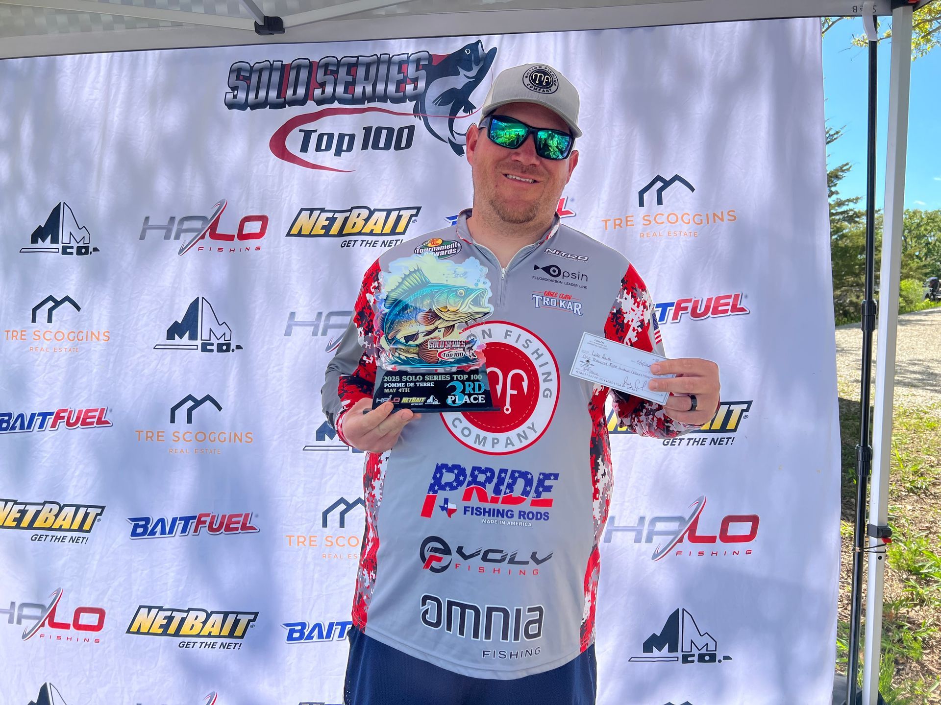 Man holding a trophy on a sponsor backdrop, wearing sunglasses and a white racing shirt.
