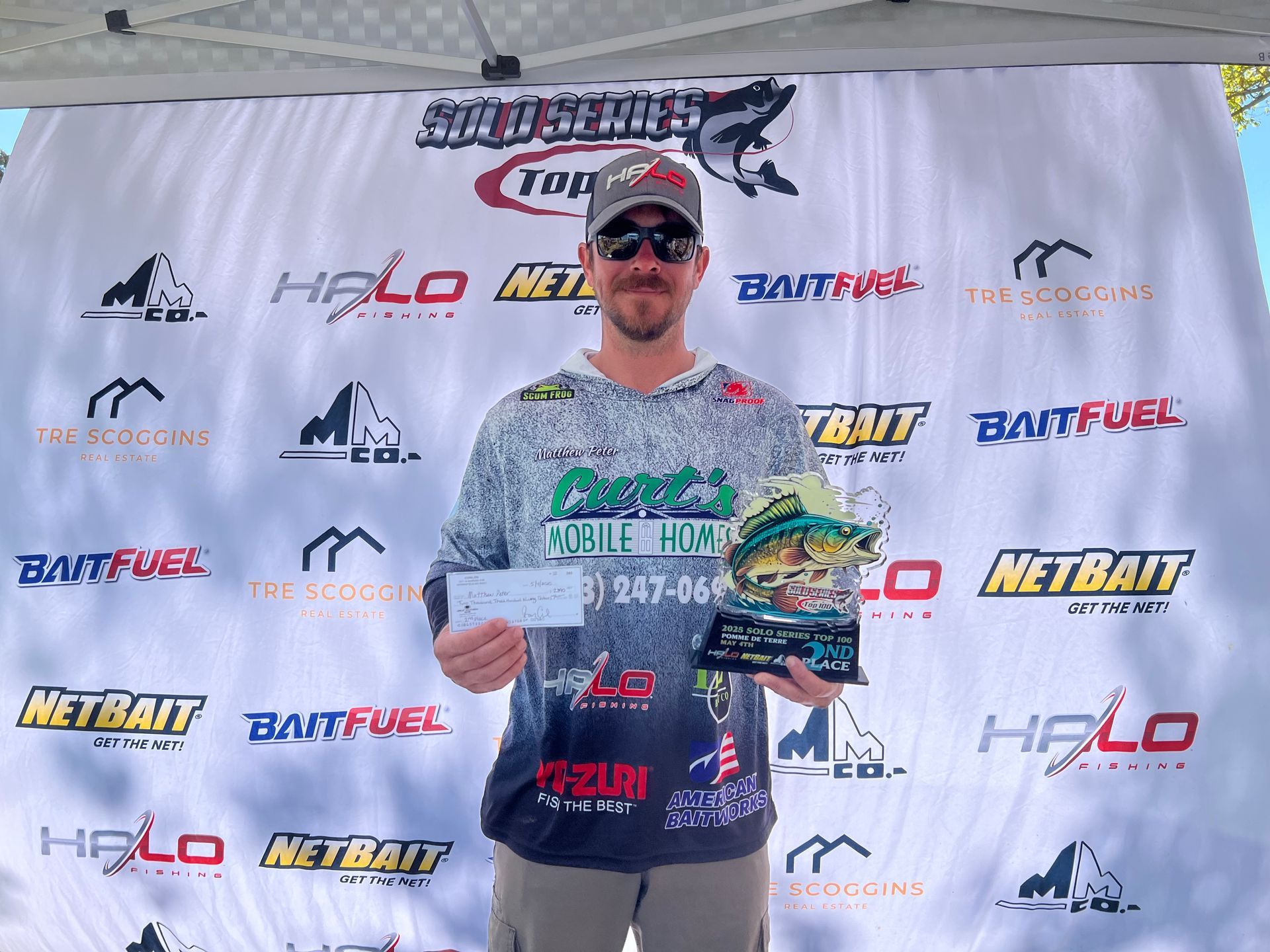 Man posing with a fishing trophy on a sponsor-branded stage, holding a check and fish-shaped award.