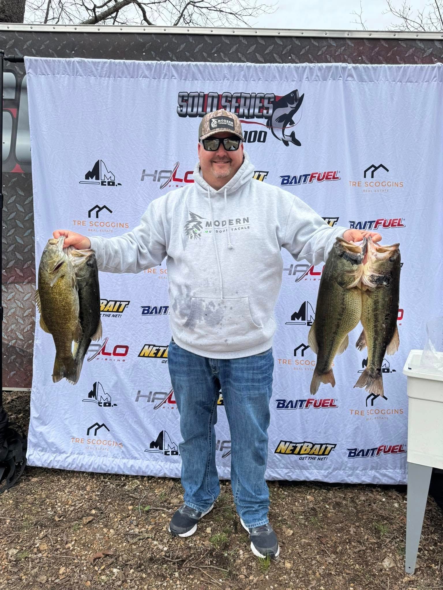 Man holding two large fish at an outdoor fishing event backdrop