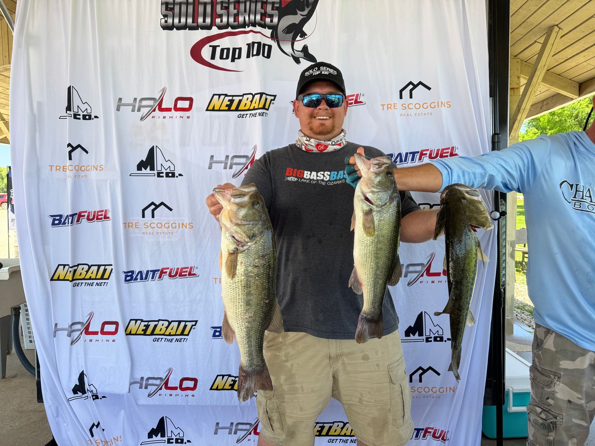 Man holding two large fish at an outdoor fishing event backdrop, wearing a black shirt and tan pants