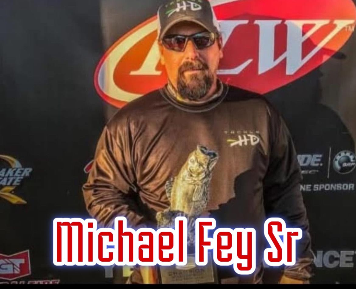 Man in black cap and hoodie holding a fish trophy before a sponsor backdrop, labeled Michael Fey Sr.