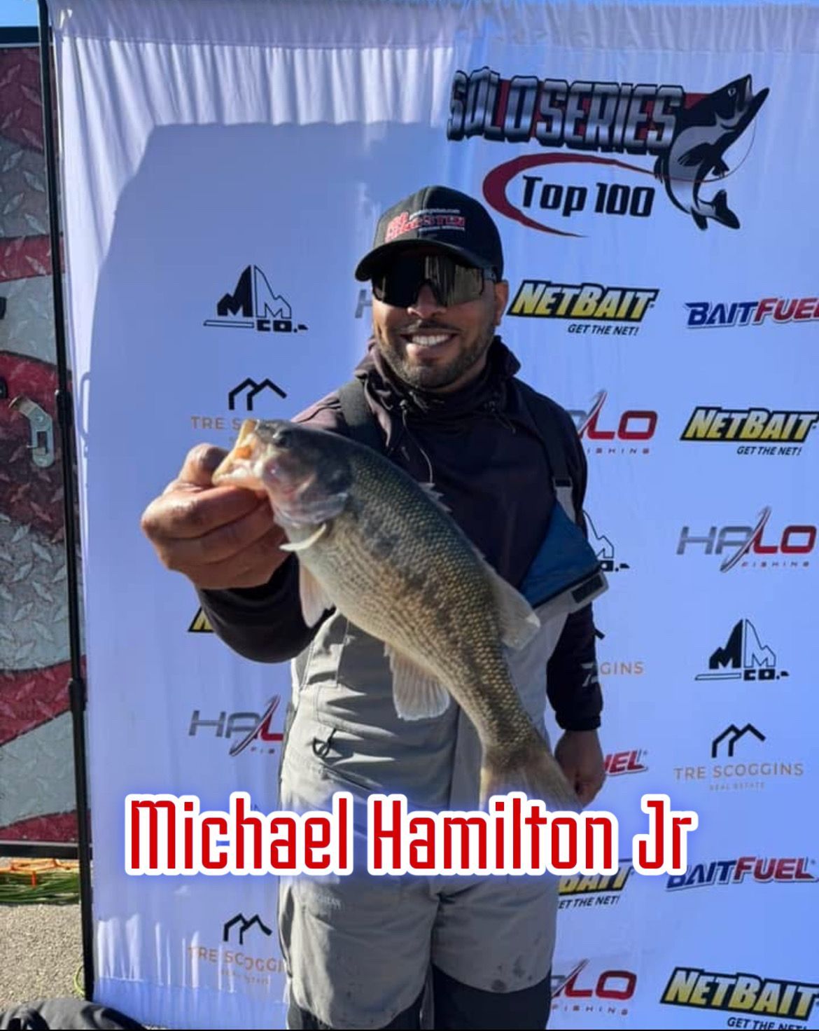 Man holding a large fish at a bass fishing event podium, smiling before sponsor backdrop