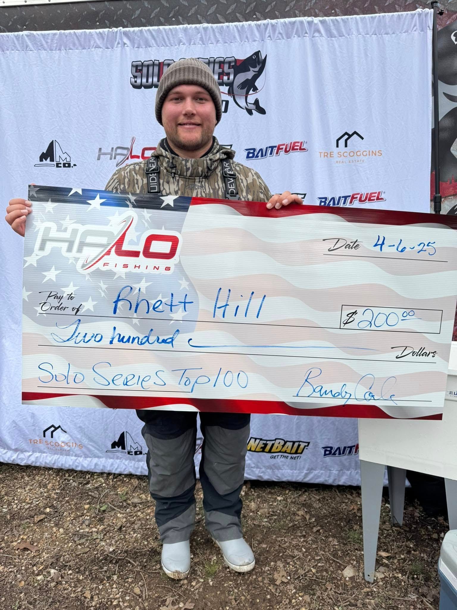 Person holding a giant signed check at an outdoor event, standing in front of a sponsor backdrop.