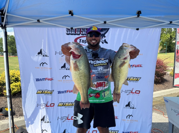 Angler holding two large fish at a bass fishing event under a sponsor banner.
