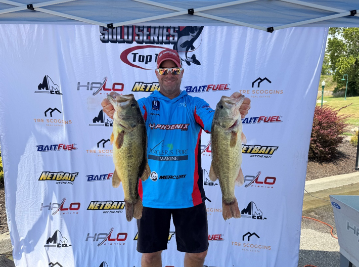 Man holding two large fish in front of a fishing tournament banner under a canopy