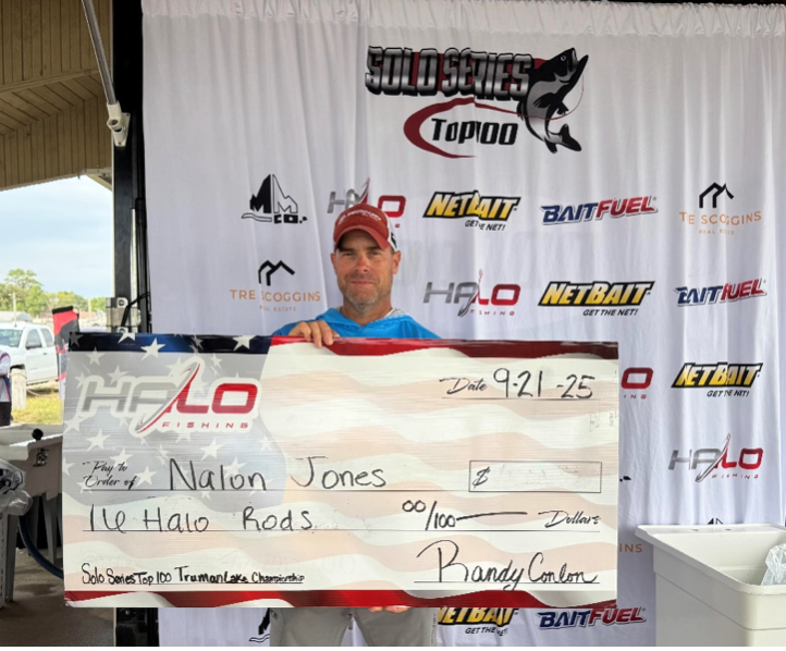 Man holding a large check at an outdoor event bannered with sponsor logos, smiling at the camera.