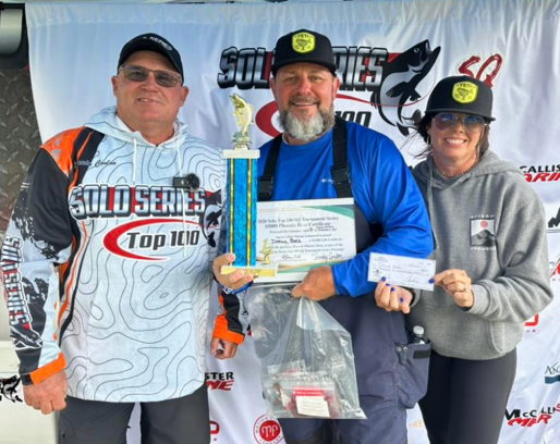Three men at a fishing event holding a trophy, certificate, and prize bags, smiling in front of a banner