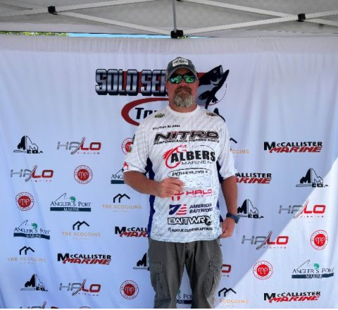 Man standing on a sponsor backdrop, wearing a white fishing jersey and cap, holding a trophy plaque.
