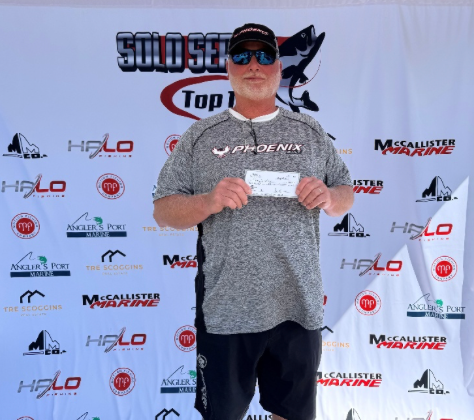 Man holding a check on a tournament stage with sponsor backdrop, wearing a gray shirt, cap, and sunglasses