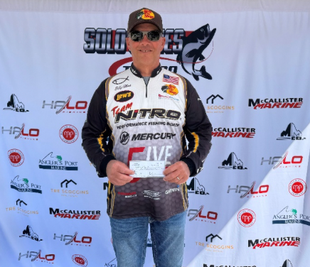 Angler holding a trophy and check at a fishing tournament podium, wearing a sponsor jersey and cap.