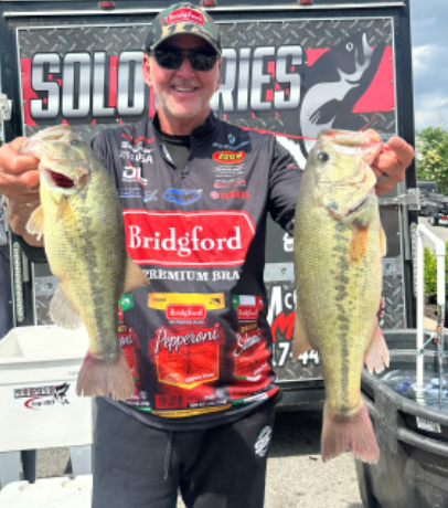 Man smiling on a boat hold two largemouth bass, standing before a “SOLD SERIES” fishing banner.