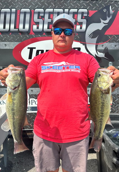 Man holding two large fish in front of a tournament fishing backdrop, wearing a red shirt and blue cap.