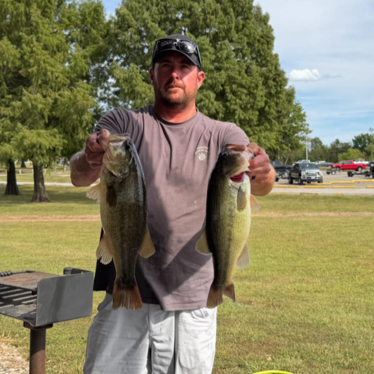 Man holding two large fish in a grassy park beside a grill, with trees and parked cars in the background.