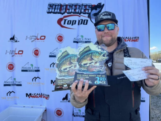 Man in sunglasses holding a fish trophy and check at a tournament podium backdrop