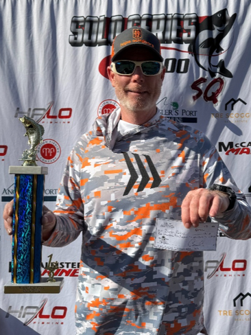 Man in orange camo shirt and cap holding a tall trophy at a sponsor backdrop