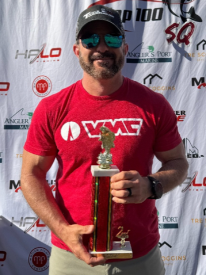 Man in a red shirt and sunglasses holding a trophy in front of a sponsor backdrop.